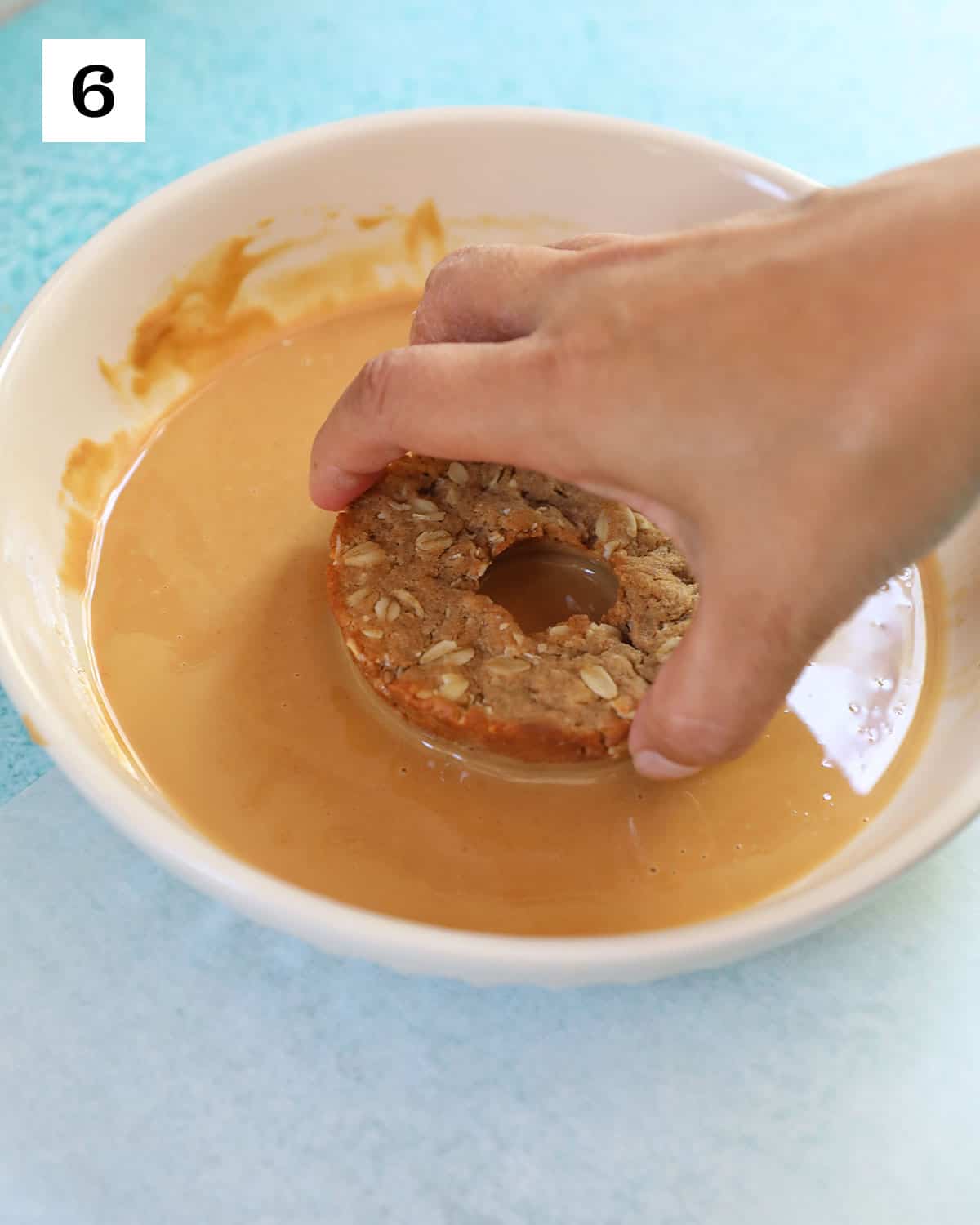 hand dipping a dog donut into peanut butter glaze in a big bowl.