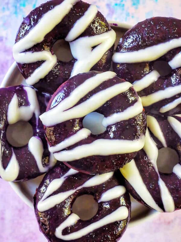 several ube donuts with ube glaze and white glaze drizzle on a purple and blue table.