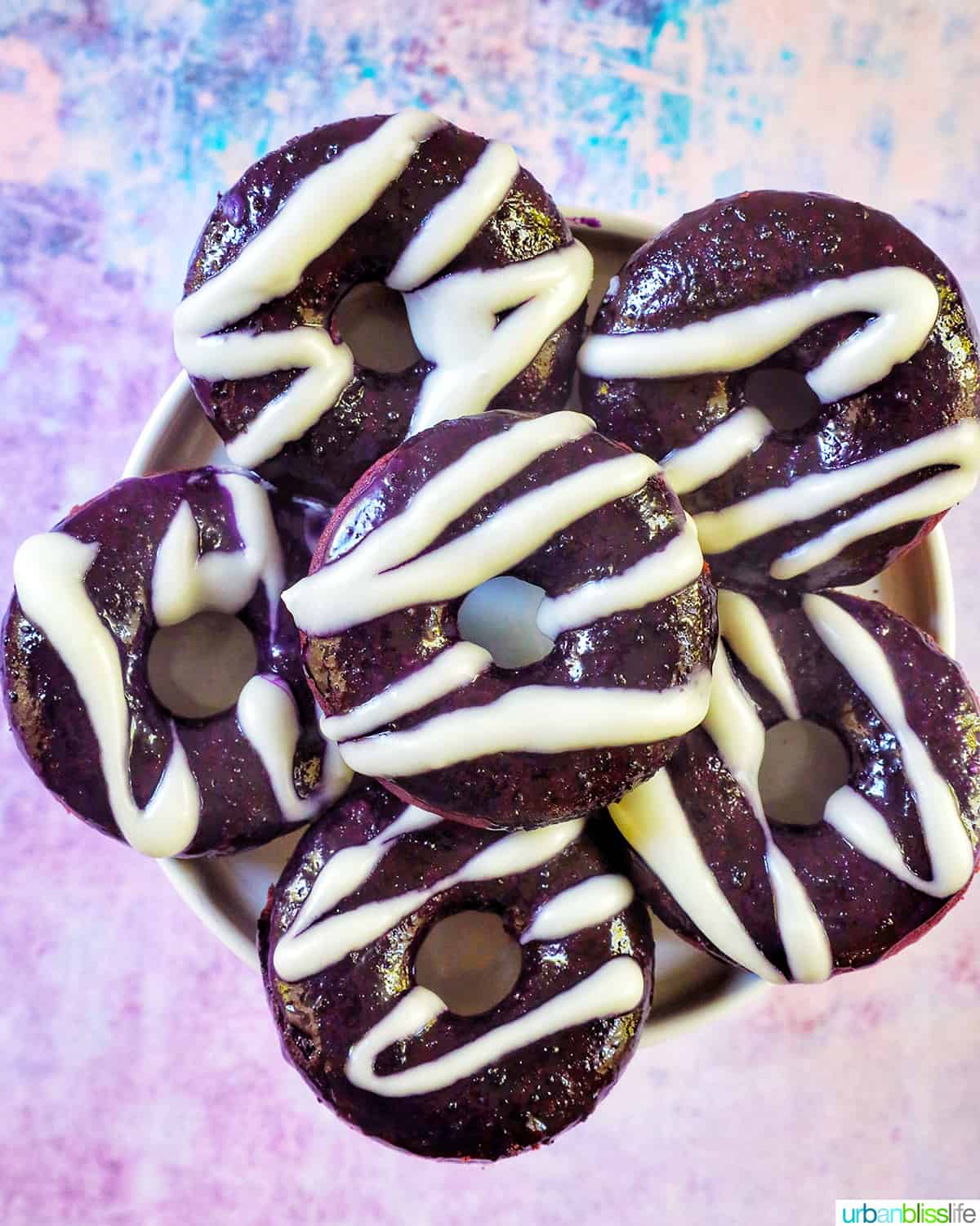 several ube donuts with ube glaze and white glaze drizzle on a purple and blue background.