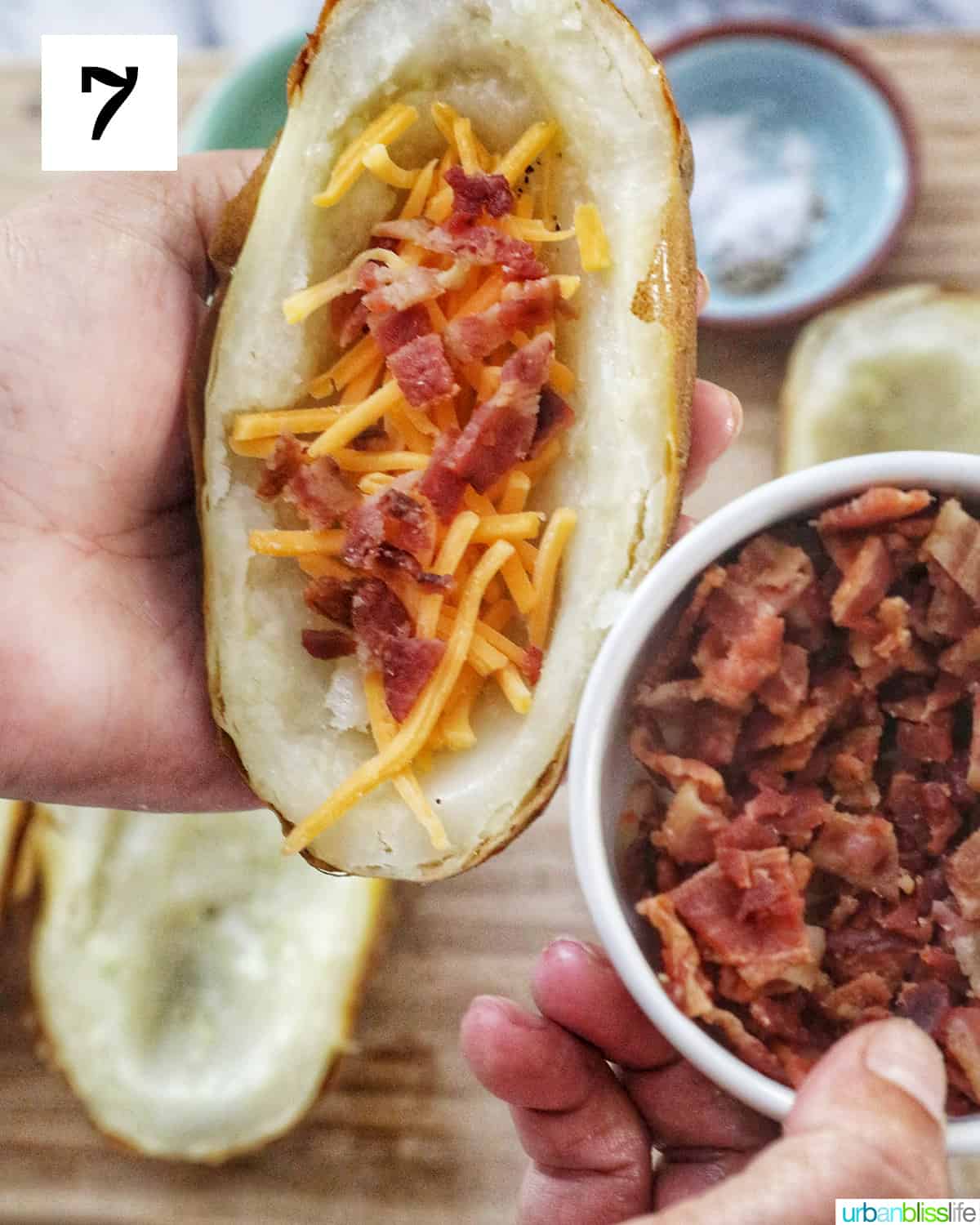 hand holding a bowl of bacon on the right and a hand holding a potato skin half topped with cheddar cheese and bacon on the left.