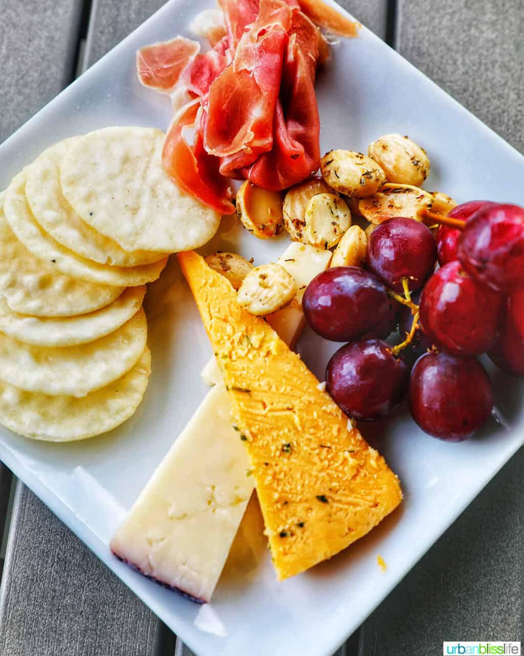 cheese and fruit plate with cheese, crackers, prosciutto, grapes, and nuts.