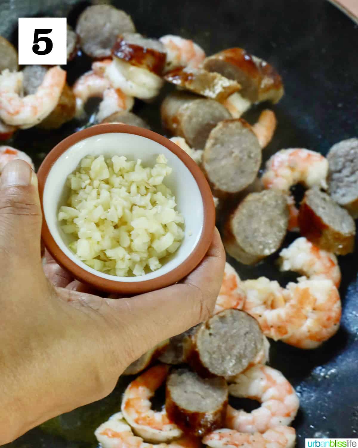 hand holding a bowl of minced garlic over a skillet of shrimp and sausage.