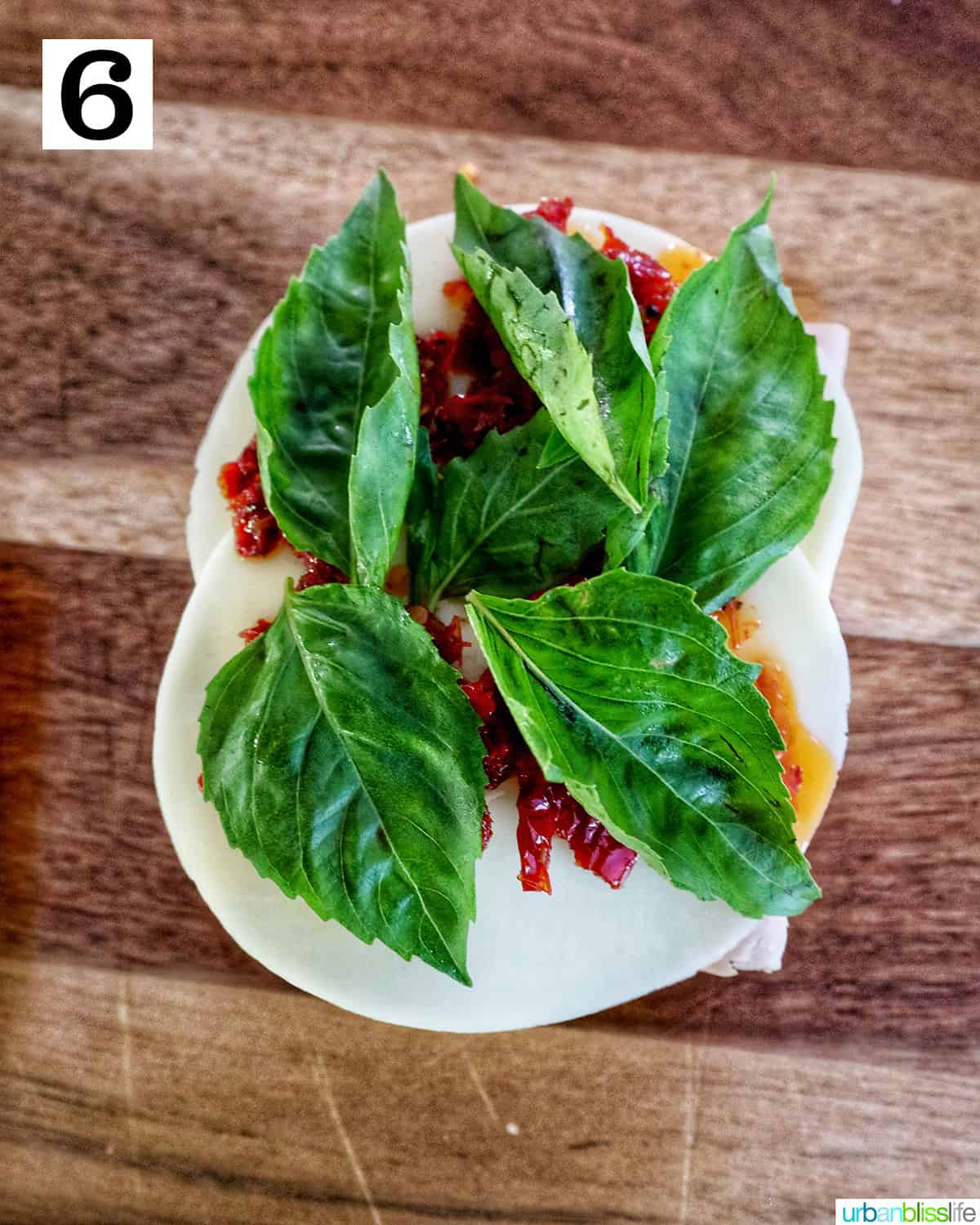basil leaves on top of sundried tomatoes and provolone cheese on a cutting board.