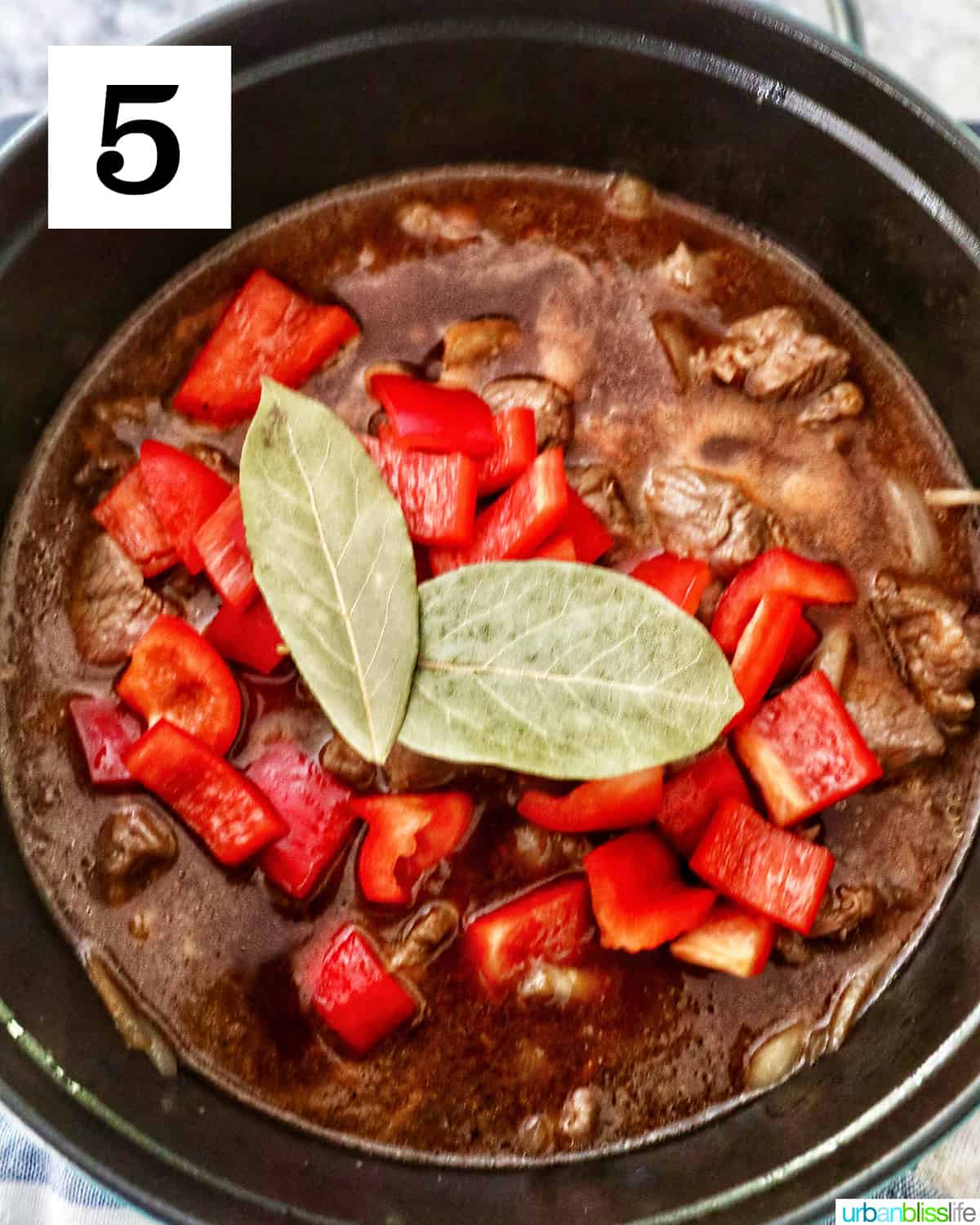 adding red bell peppers and two bay leaves to beef mechado in a dutch oven.