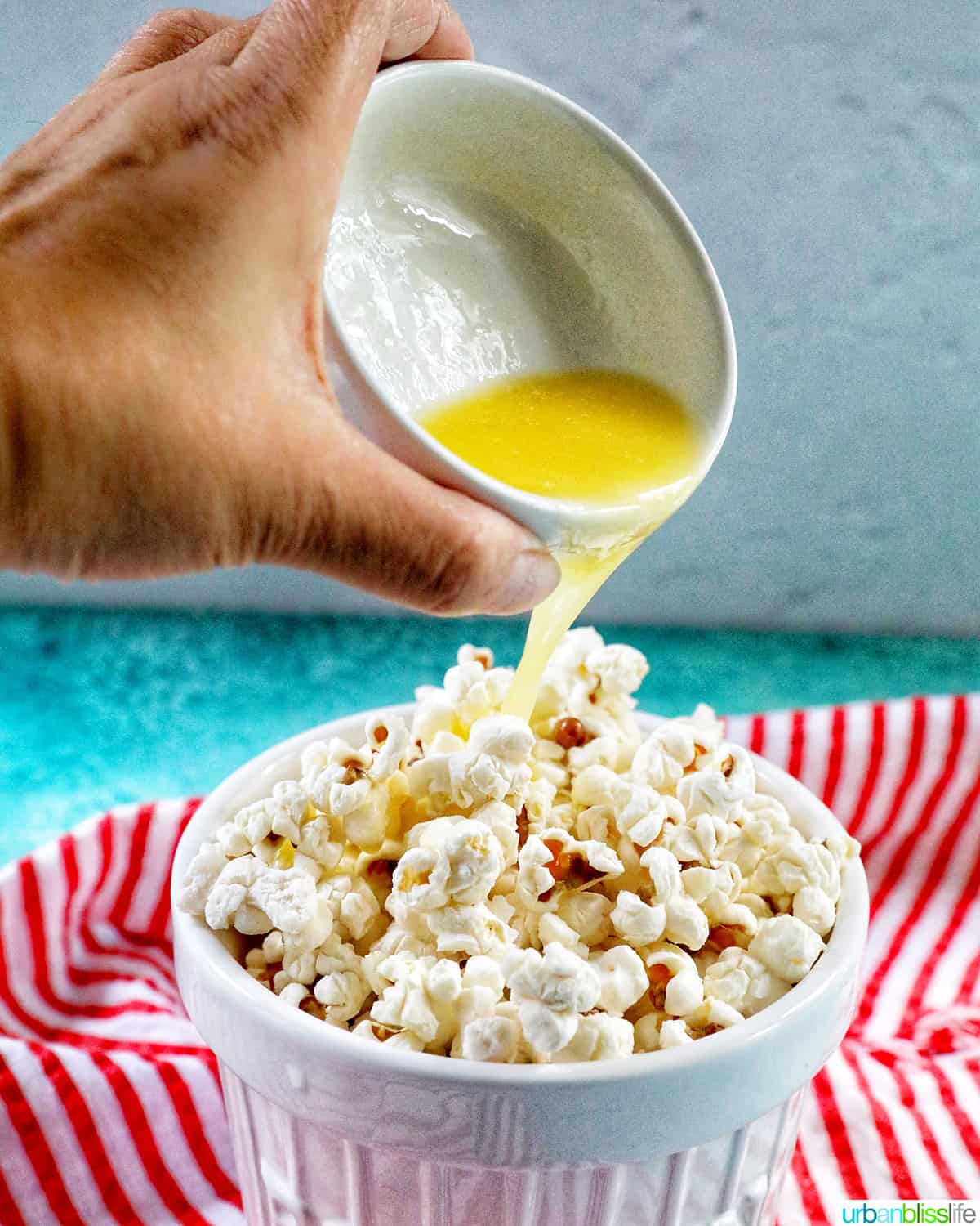 hand pouring a bowl of melted butter over white popcorn bowl full of Instant Pot Popcorn on a red and white napkin.