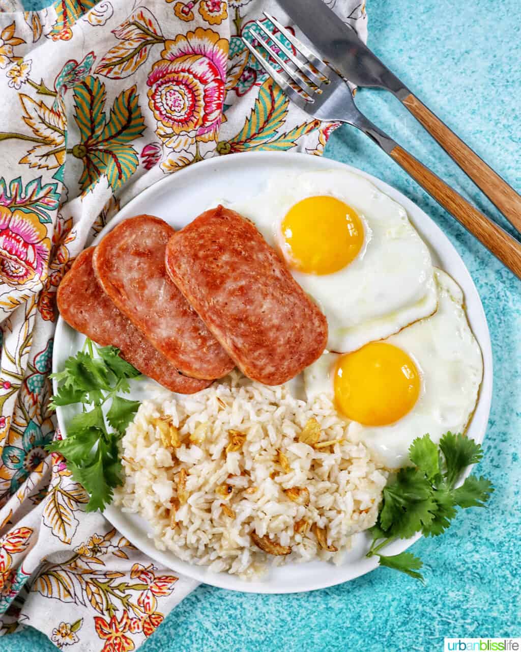 Plate of Spamsilog: slices of Spam, two eggs over easy, and Filipino Sinangag garlic rice with greens and fork and knife.