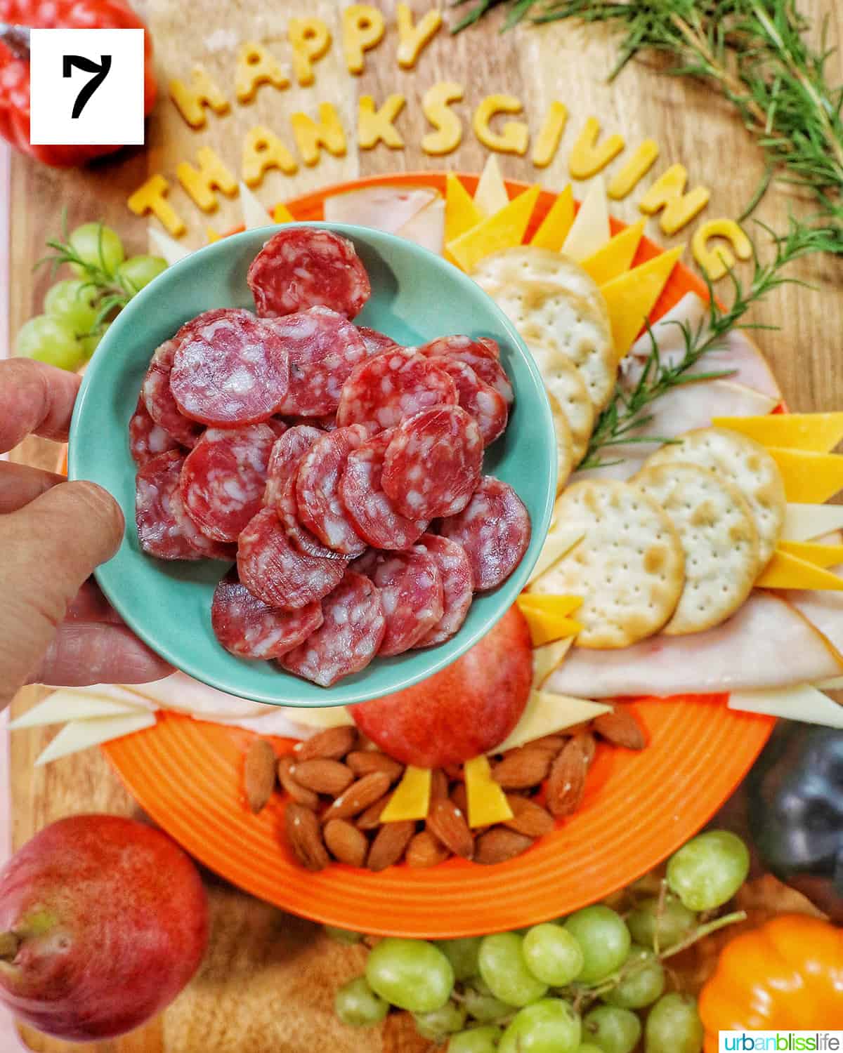hand holding a plate of sliced salami over a Thanksgiving charcuterie board.