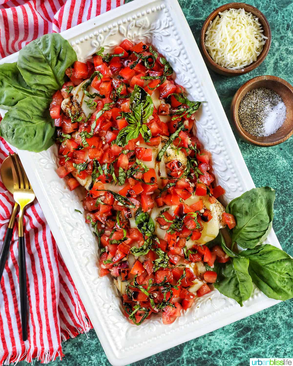 chicken bruschetta with basil leaves in a large white serving platter, with bowl of cheese, utensils on red and white striped napkin, on a green table.