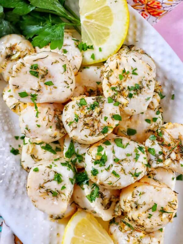 several cooked scallops topped with herbs and garlic, with lemon slices and parsley, on a white plate.