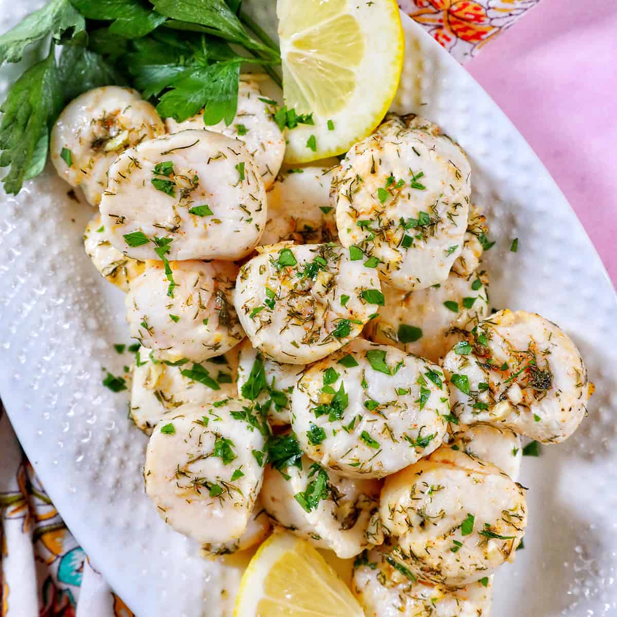 several cooked scallops topped with herbs and garlic, with lemon slices and parsley, on a white plate.