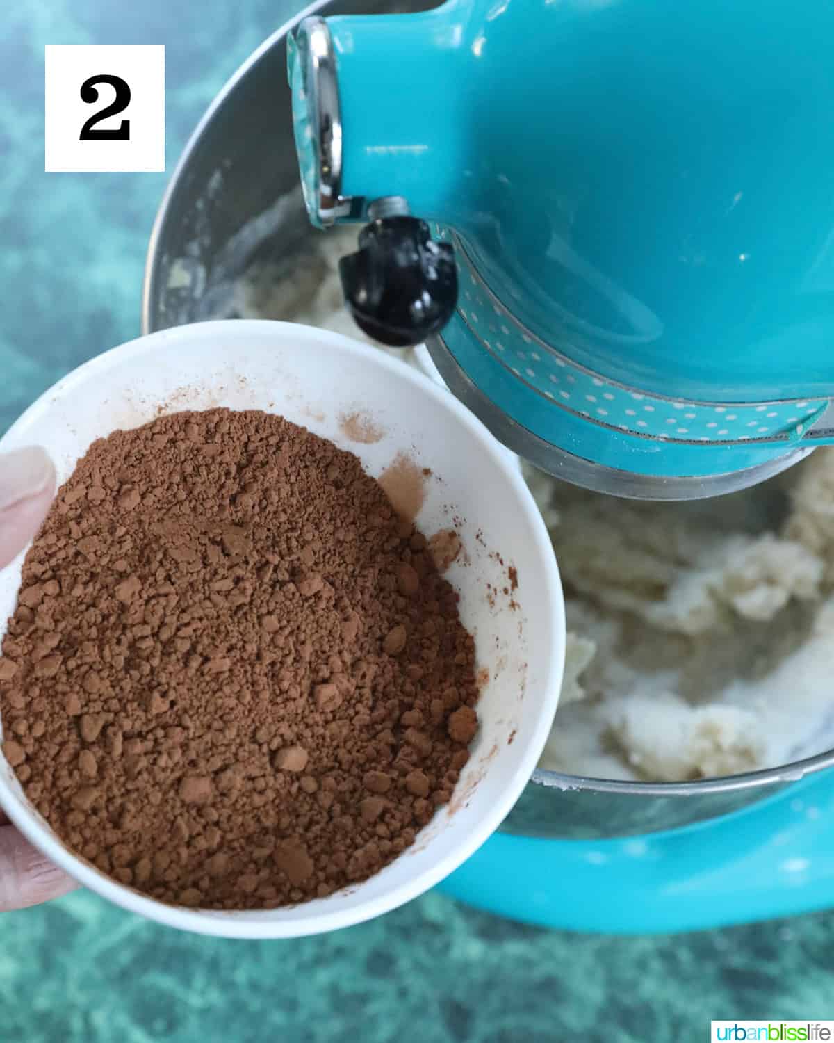 bowl of cocoa powder being added to a blue stand mixer to make chocolate buttercream frosting.