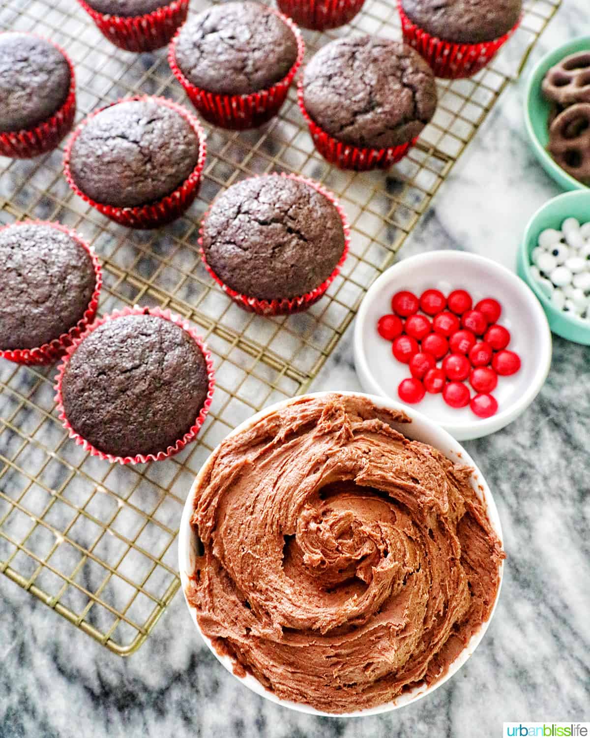 bowl of chocolate buttercream frosting next to several chocolate cupcakes and candy decorations.