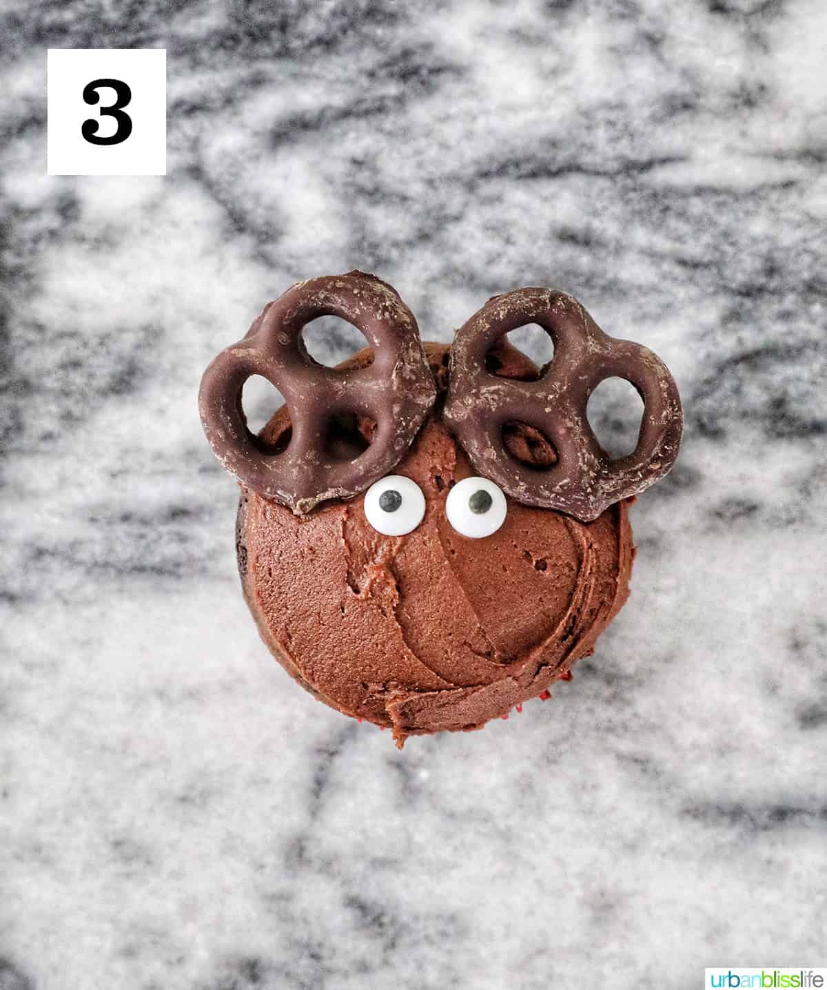 chocolate covered pretzels and candy eyes on top of a chocolate frosted cupcake on a marble counter.