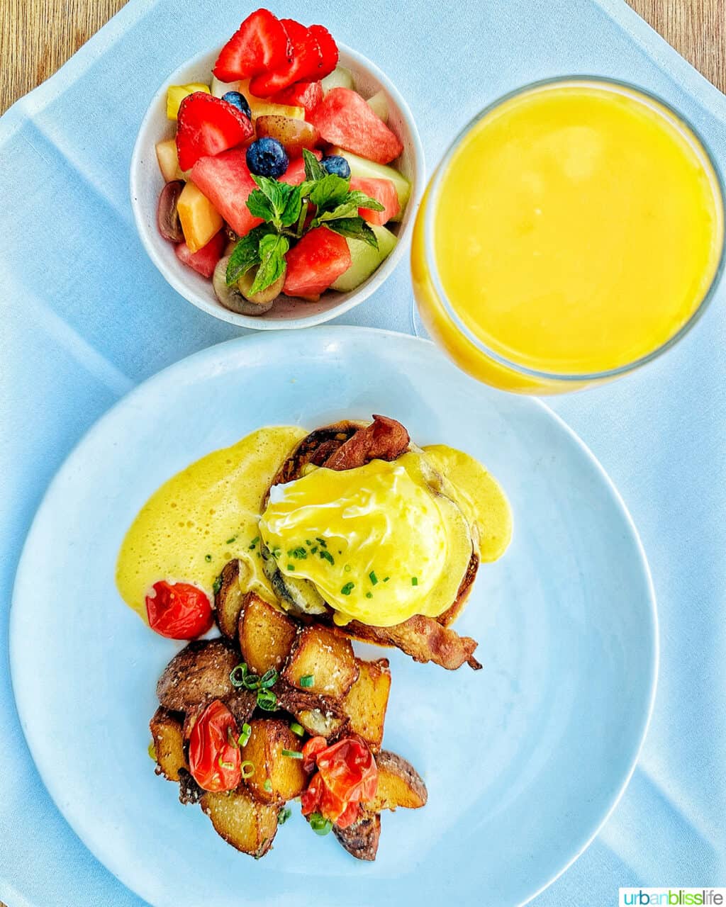 eggs benedict with potatoes on a plate, with glass of orange juice and side of fruit.