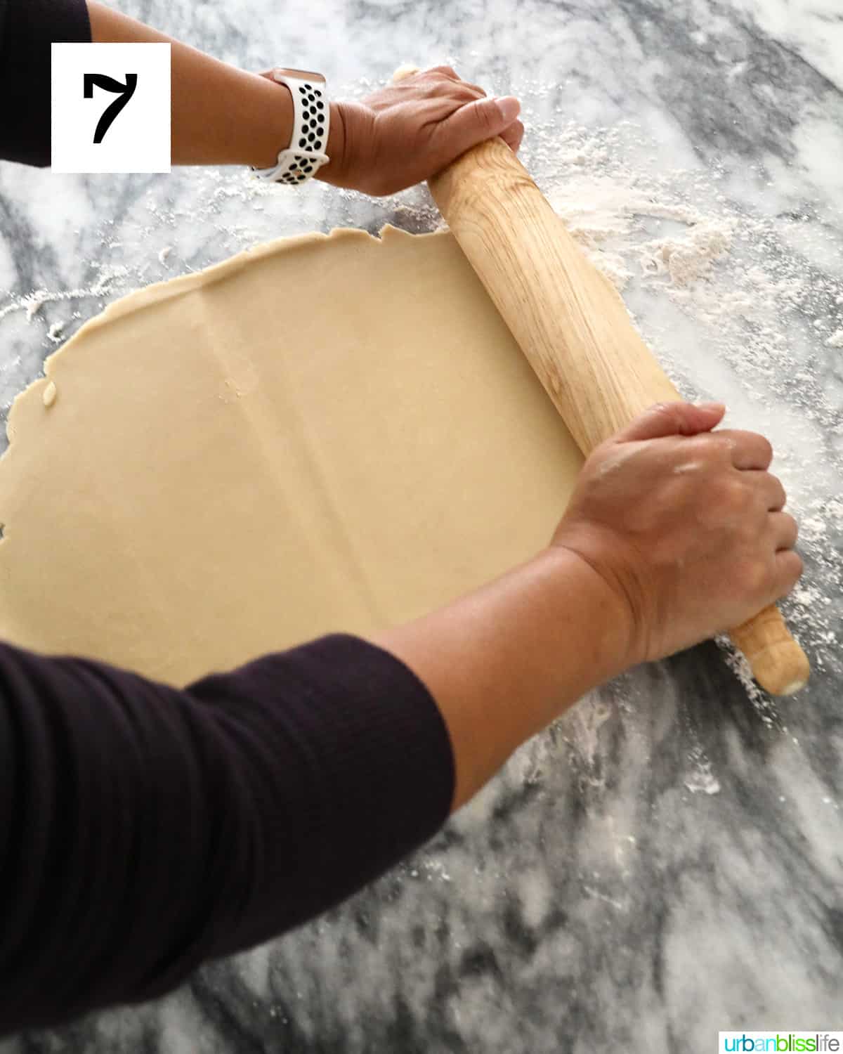 hands and arms rolling out cookie dough with a rolling pin on a marble table.