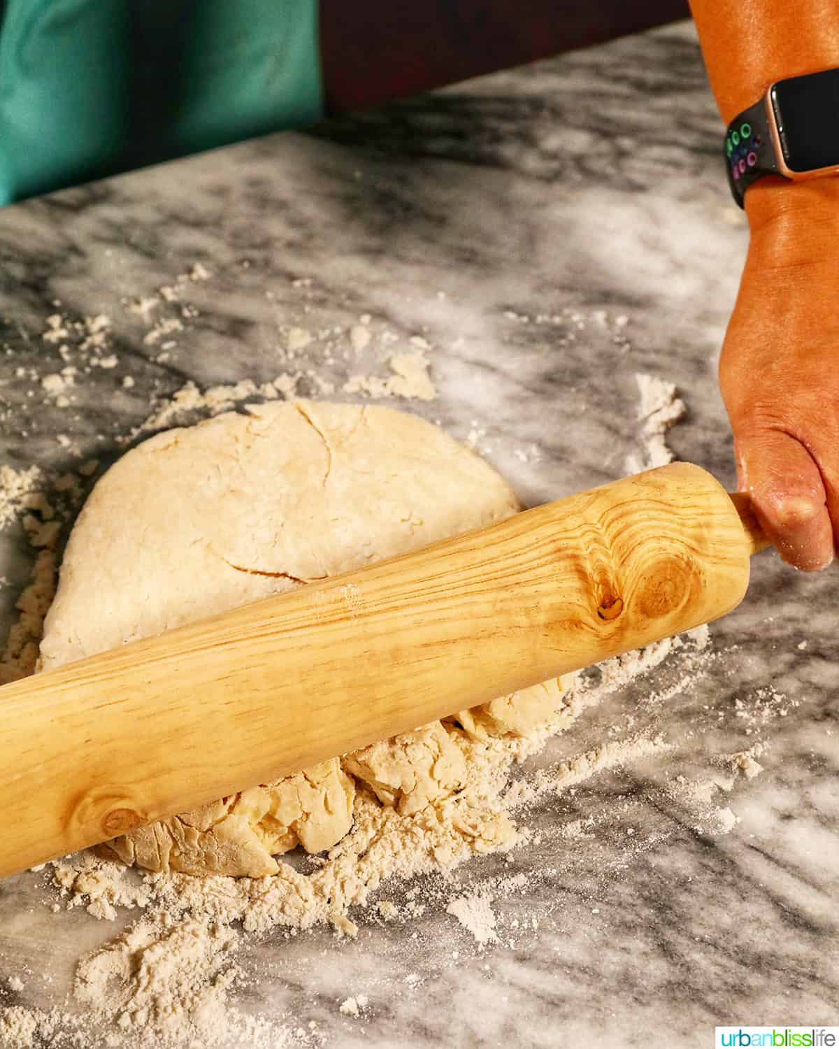 hands and rolling pin rolling biscuit dough flat on a marble counter.