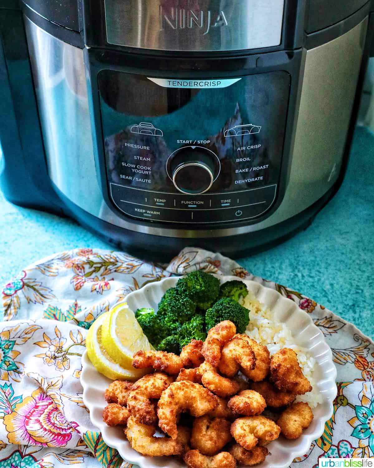 ninja foodi multicooker behind a plate of popcorn shrimp with broccoli and lemon.