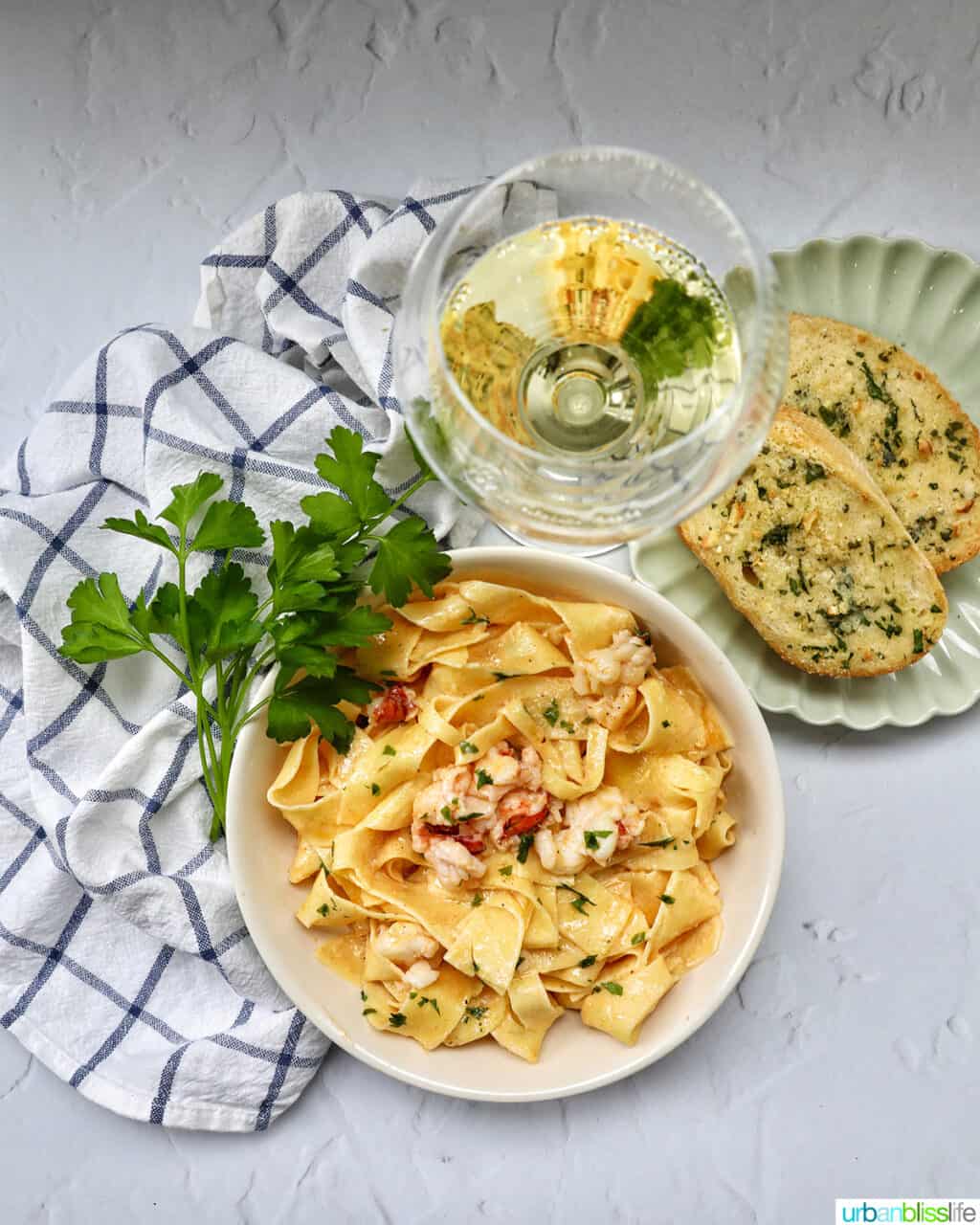 bowl of lobster pasta with side of garlic bread, herbs, glass of white wine, and a kitchen towel.