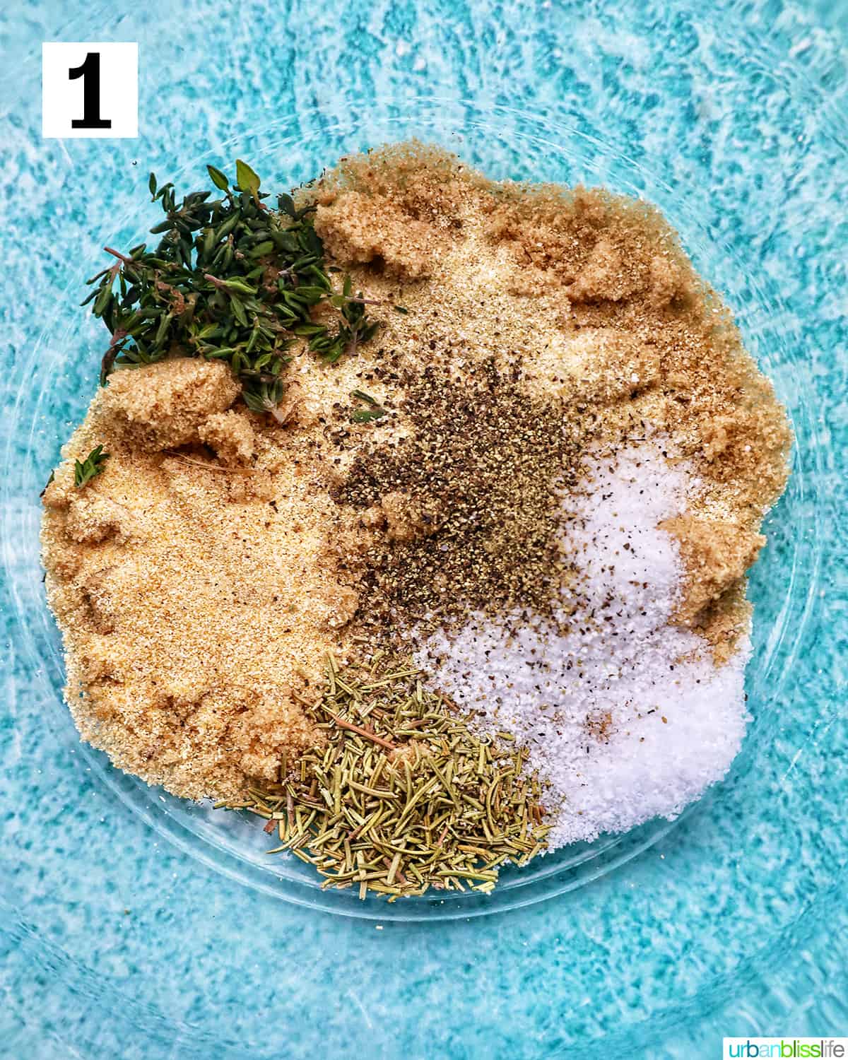 herbs and seasonings in a clear glass bowl on blue table.