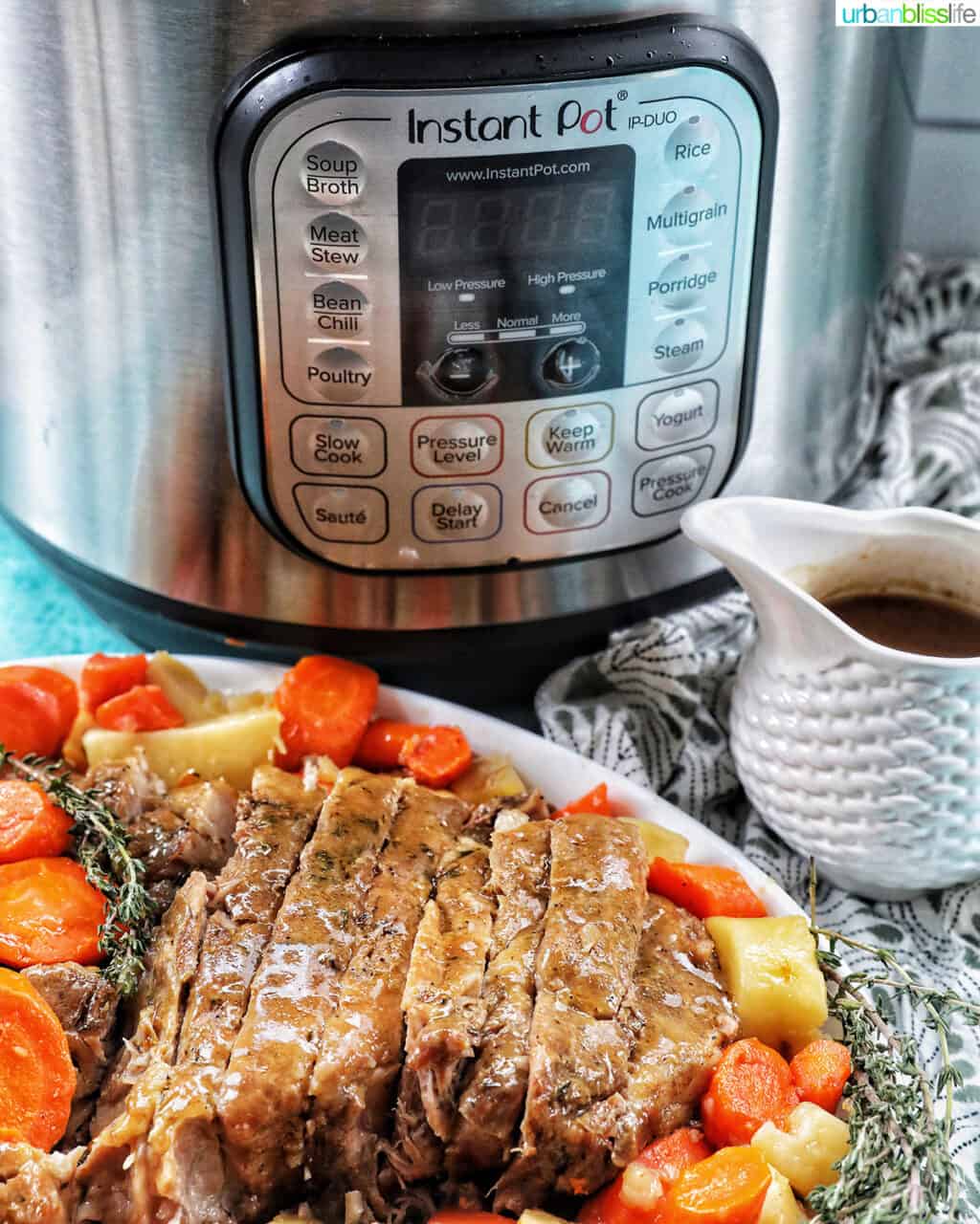 Sliced pork roast with carrots and potatoes on a plate in front of an Instant Pot and a gravy boat with gravy.