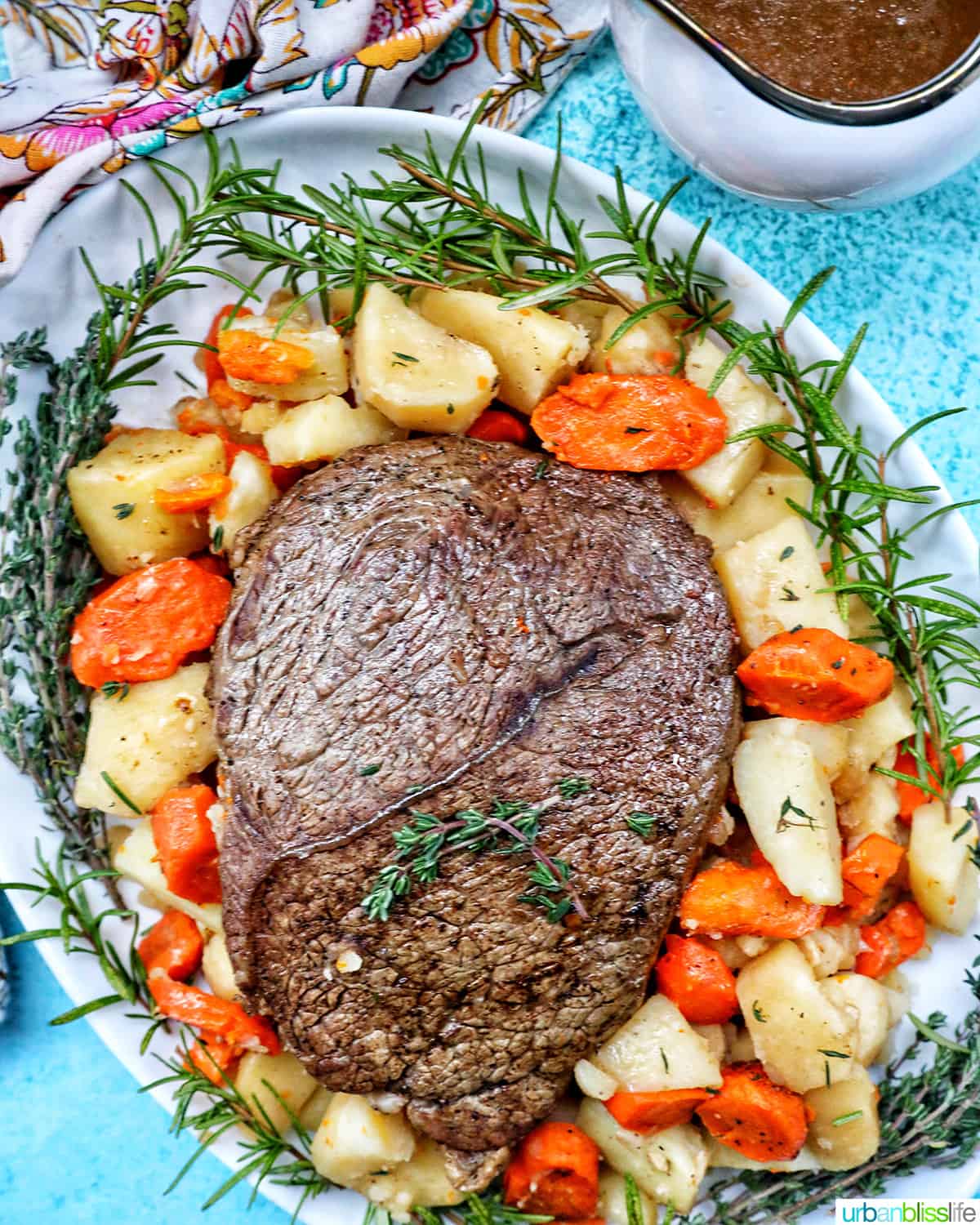 serving plate with cooked sirloin tip roast surrounded by carrots, potatoes, and herbs with a side of gravy.