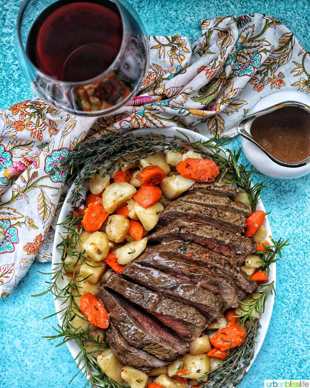 Sliced sirloin tip roast surrounded by carrots, potatoes, and herbs with a side of gravy and glass of red wine.