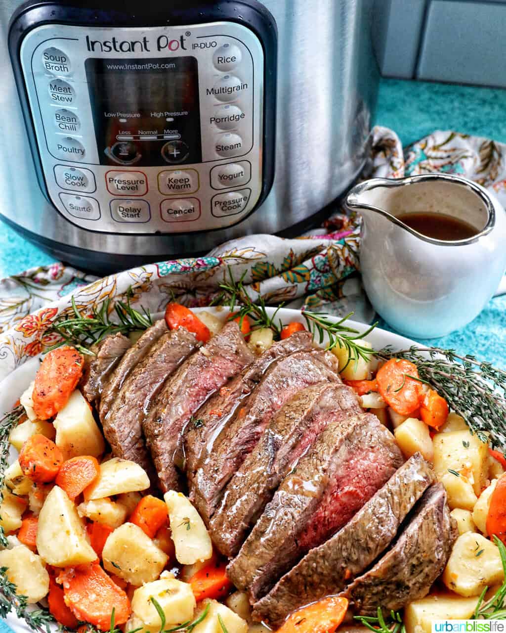 Sliced sirloin tip roast surrounded by carrots, potatoes, and herbs in front of an Instant Pot with a side of gravy.