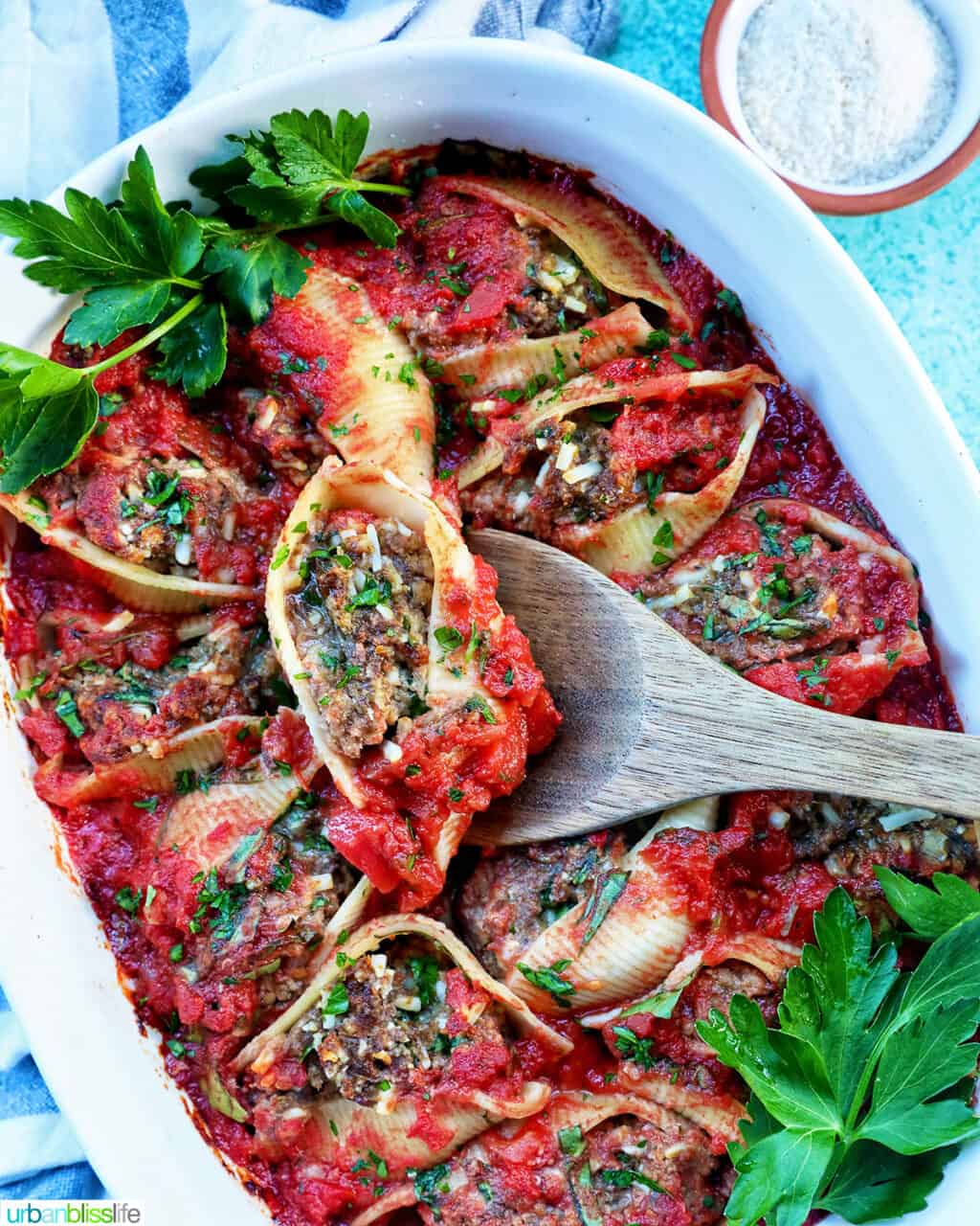 wooden spoon holding up a stuffed shells with ground beef over a casserole dish full of the pasta course.