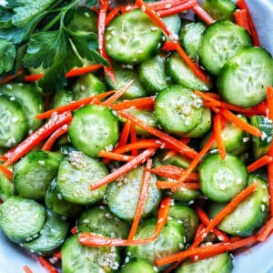 Bowl of sliced cucumbers, julienned carrots, sesame seeds, and herbs on the side.