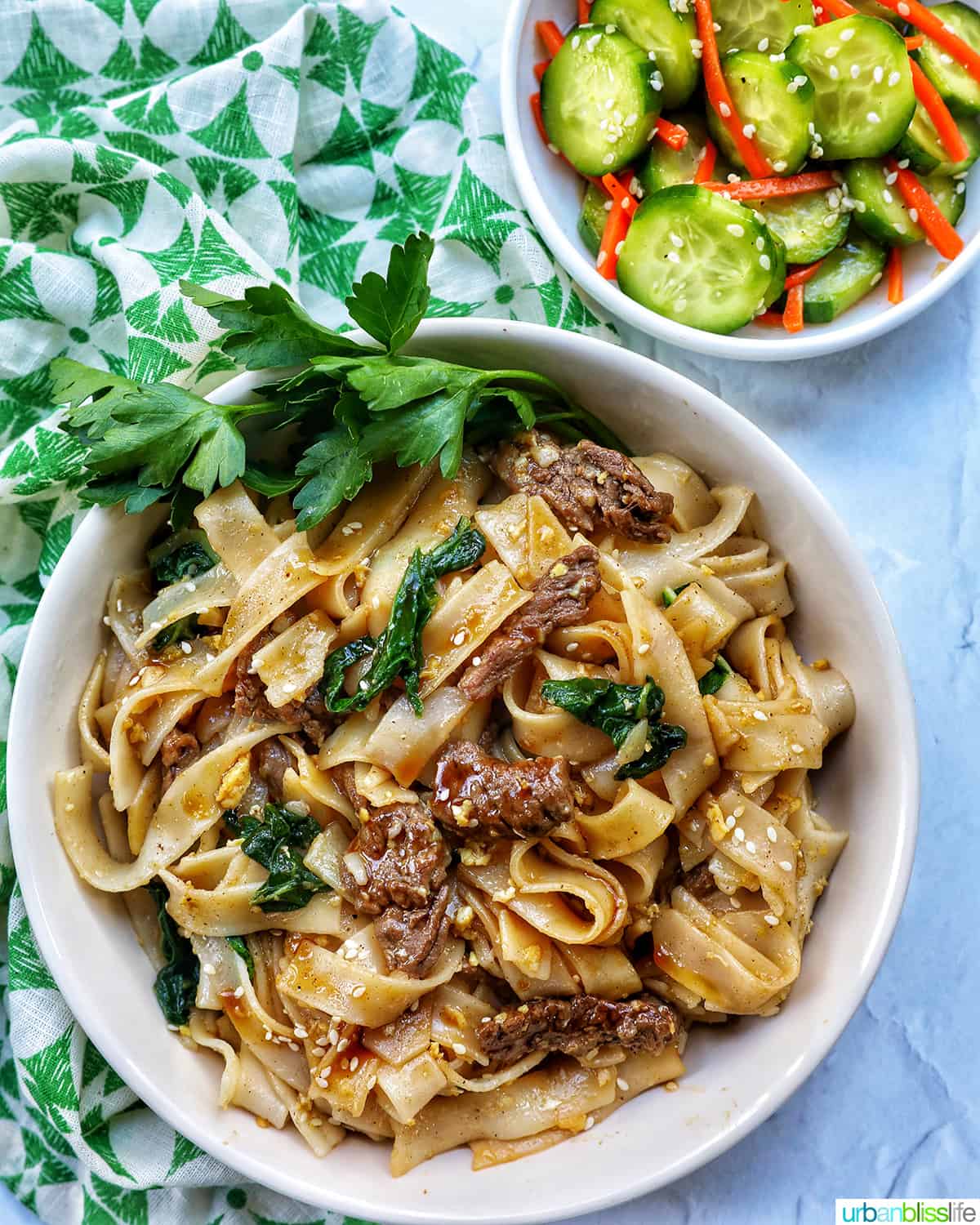 bowl of beef pad see ew noodles, bok choy, chopsticks with side of cucumber salad.