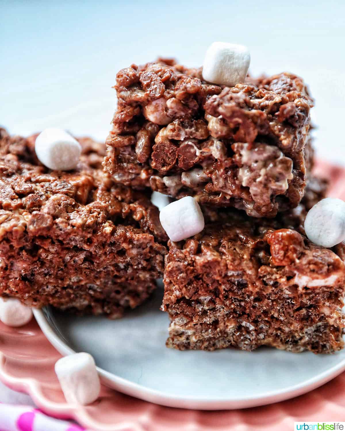 Three treats of hocolate rice krispies stacked with marshmallows on a pink and white plate.