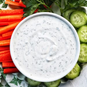 bowl of dairy free ranch dressing with carrot sticks, cucumber slices, and herbs.