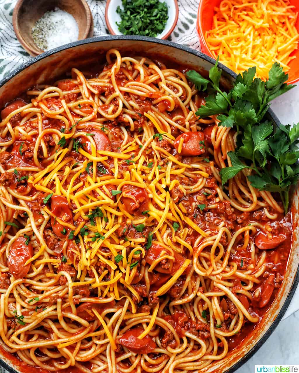 Filipino-style spaghetti in a saucepan with side of salt, pepper, parsley, shredded cheese.