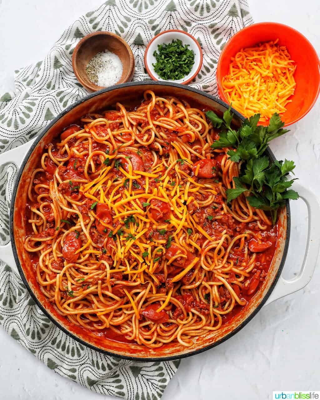 Filipino-style spaghetti in a saucepan with side of salt, pepper, parsley, shredded cheese.