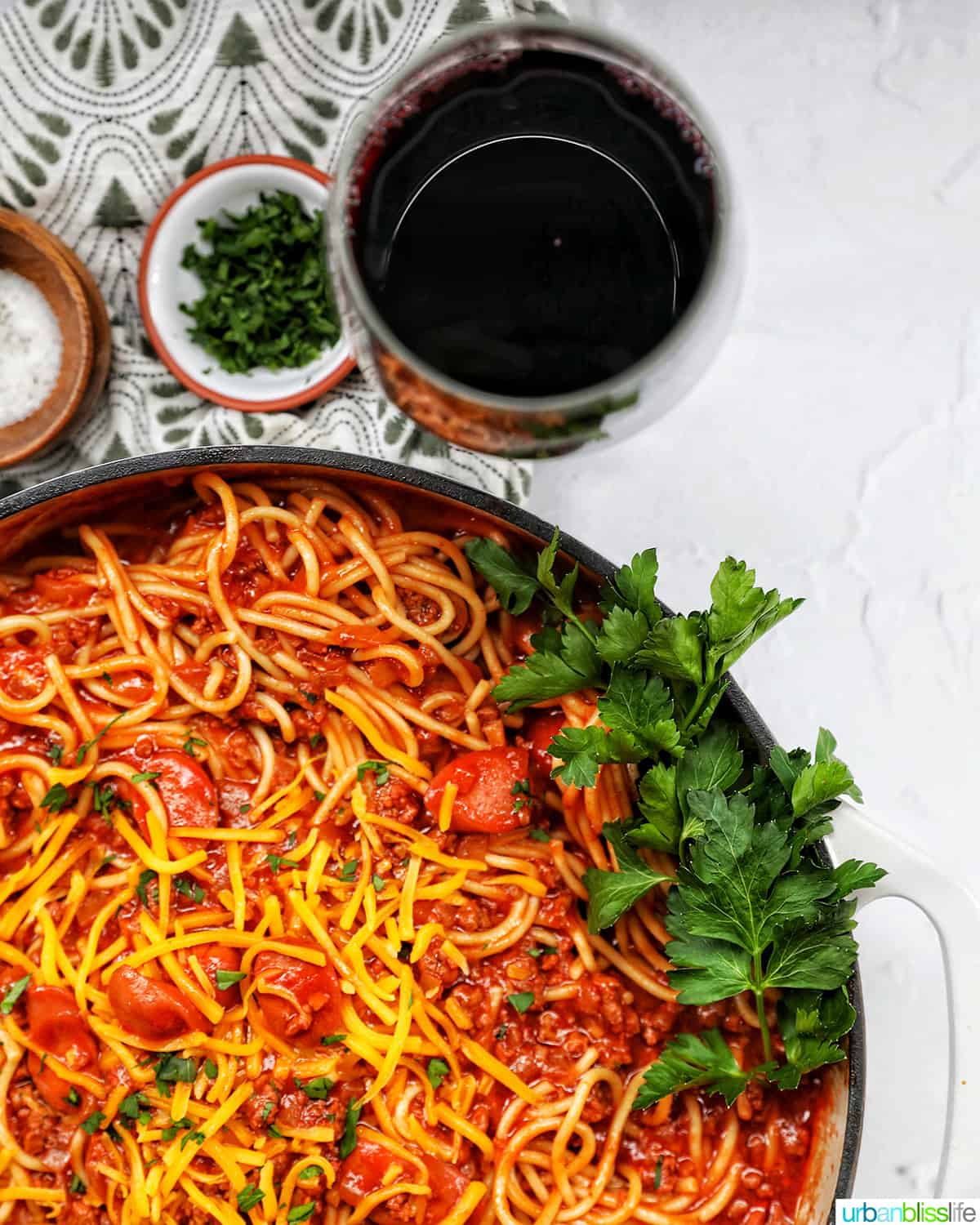 Filipino-style spaghetti in a saucepan with glass of red wine and side of salt, pepper, parsley, shredded cheese.