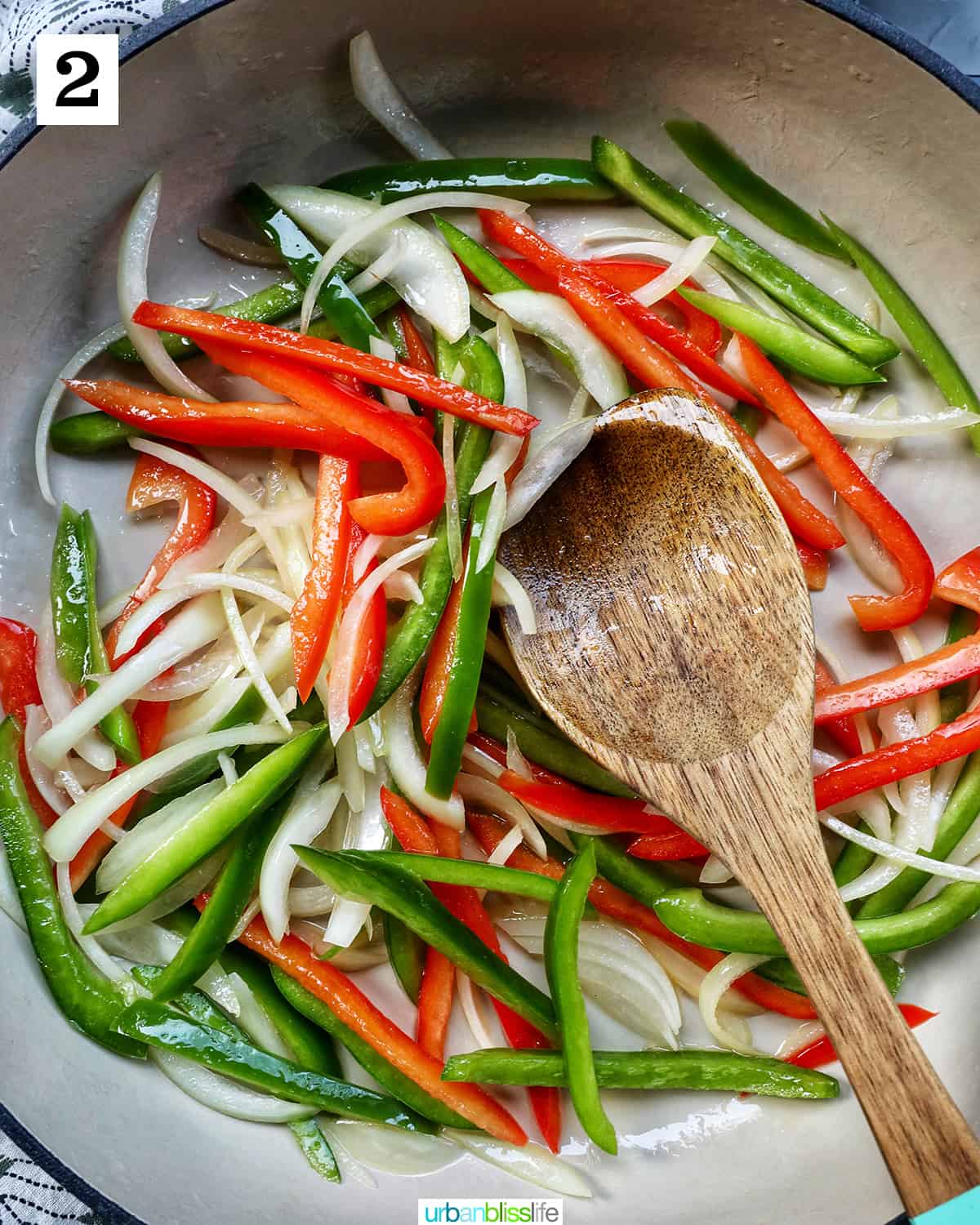 wooden spoon with sliced onions, red peppers, and green peppers in a saucepan.