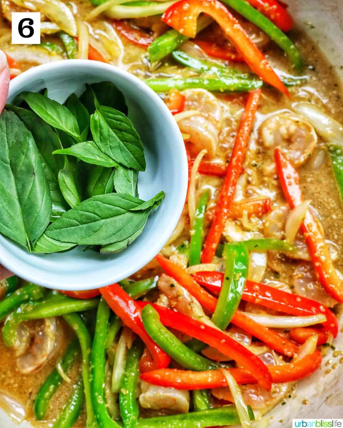 bowl of Thai basil leaves over sliced onions, red peppers, and green peppers in a saucepan.