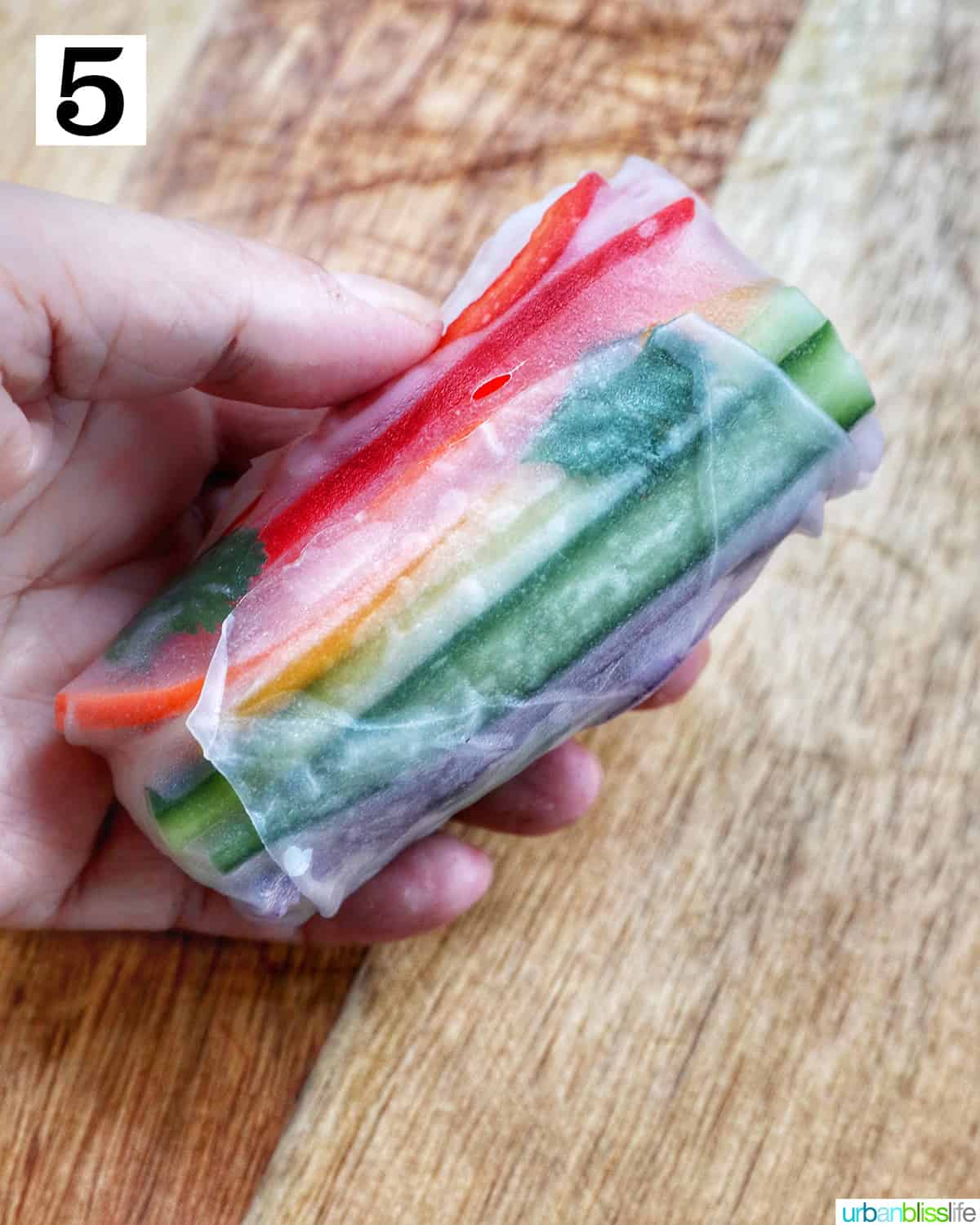 hand holding a rolled up vegan summer roll filled with sliced colorful vegetables above a wooden cutting board.