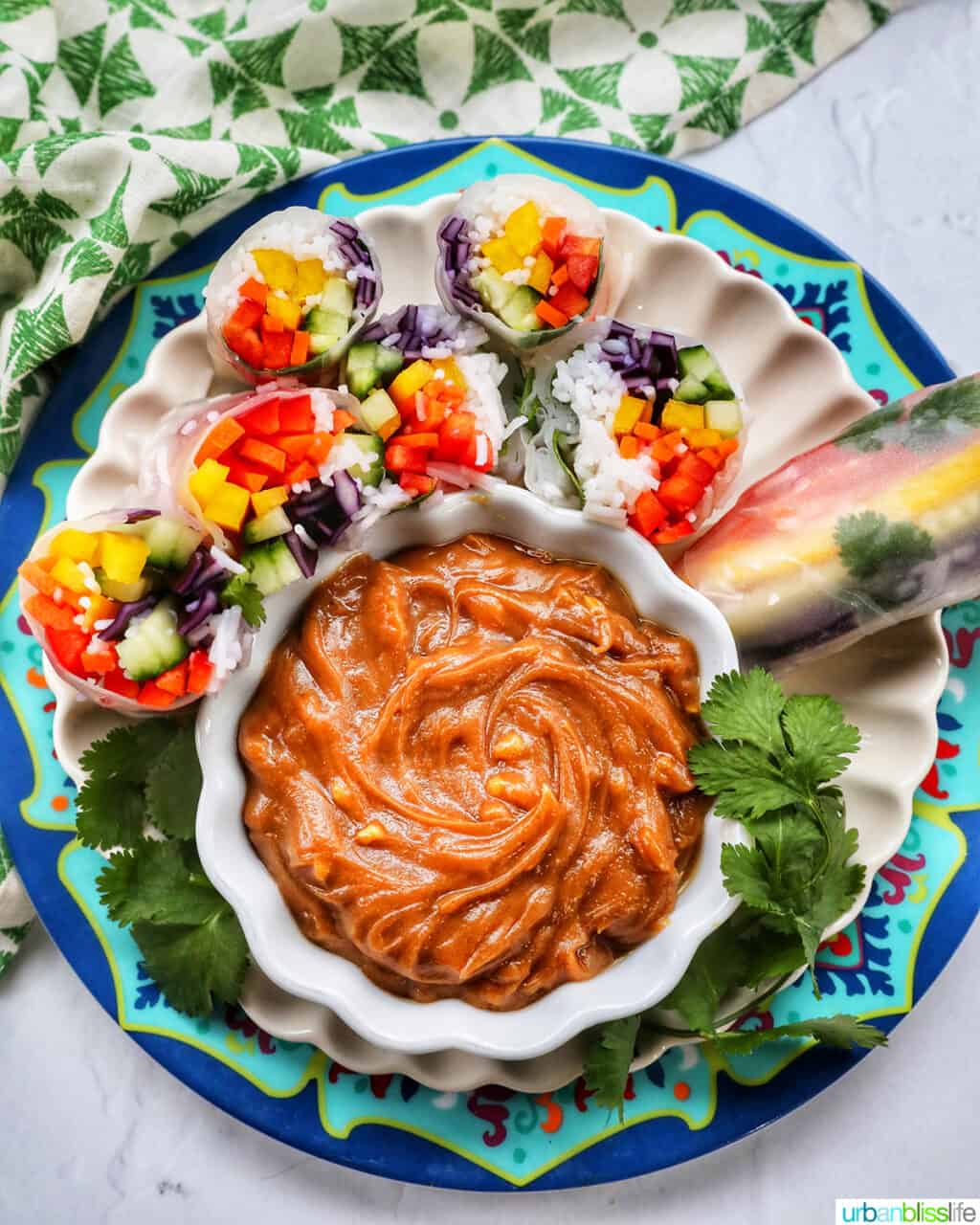 colorful plate with several vegan summer rolls cut in half showing the vegetables and vermicelli noodles with a side of thai peanut sauce in a bowl.