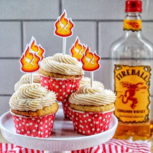 fireball cupcakes in red and white polka dot cupcake liners on a cake pedestal with buttercream frosting and fire sticker decorations on top and a bottle of Fireball in the background.