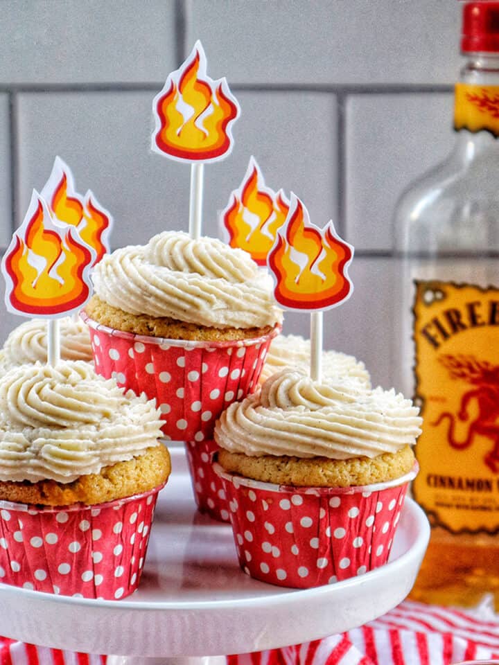 fireball cupcakes in red and white polka dot cupcake liners on a cake pedestal with buttercream frosting and fire sticker decorations on top and a bottle of Fireball in the background.