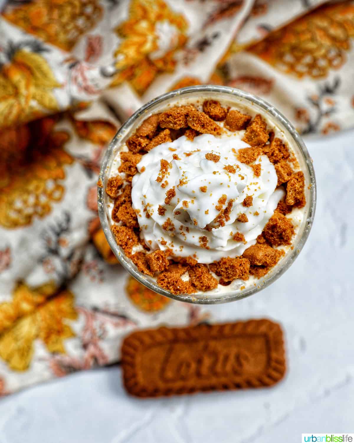 overhead photo of Biscoff Milkshake in a milkshake glass with a yellow floral kitchen linen and biscoff cookie.