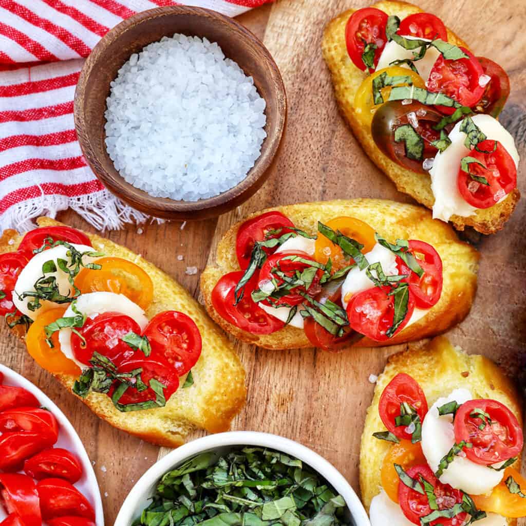 crostini with tomatoes, mozzarella, basil surrounded by plates of the same toppings and a bowl of sea salt.