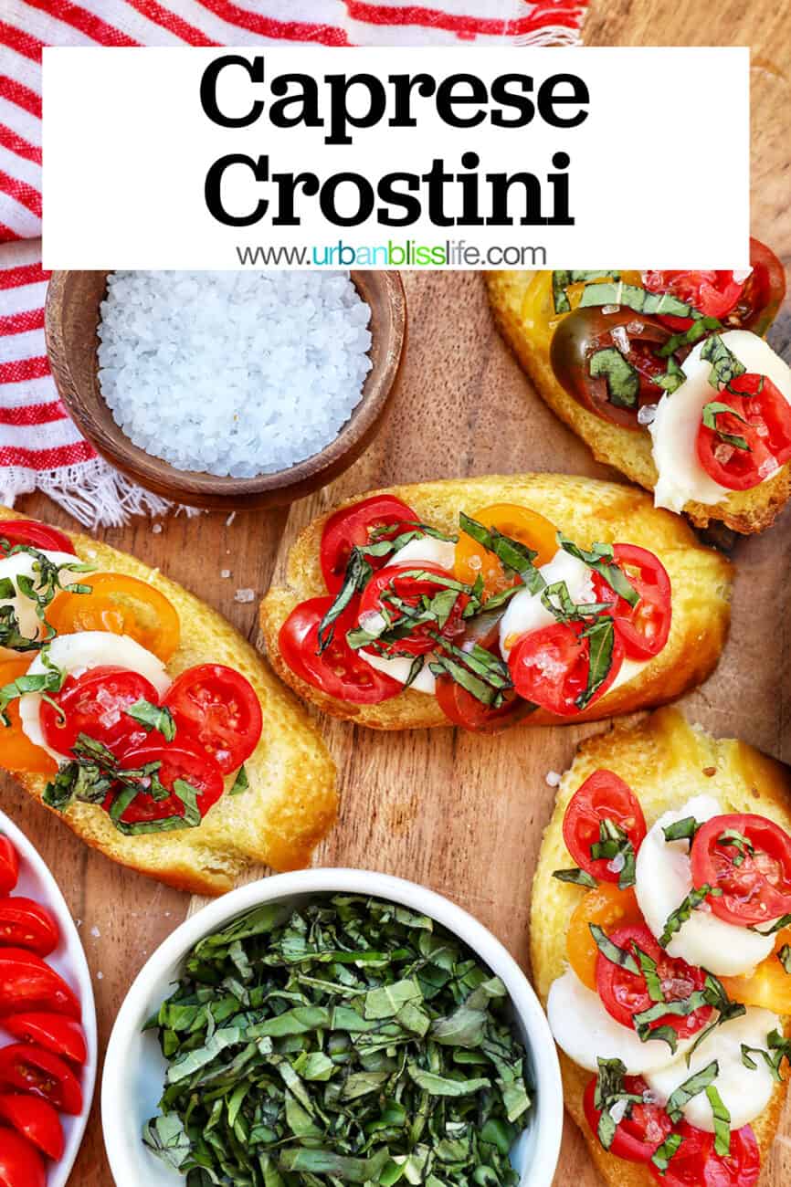 crostini with tomatoes, mozzarella, basil surrounded by plates of the same toppings and a bowl of sea salt with title text that reads Caprese Crostini.