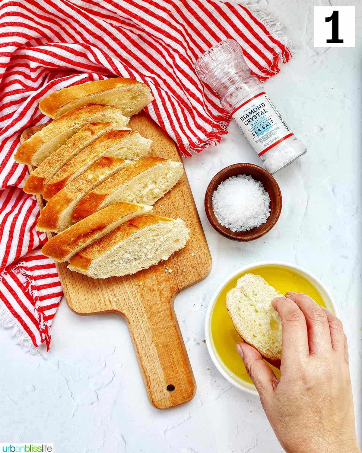 hand dipping a crostini into a bowl of olive oil with crostini slices on a wooden cutting board on red and white towell with bowl and shaker of sea salt.