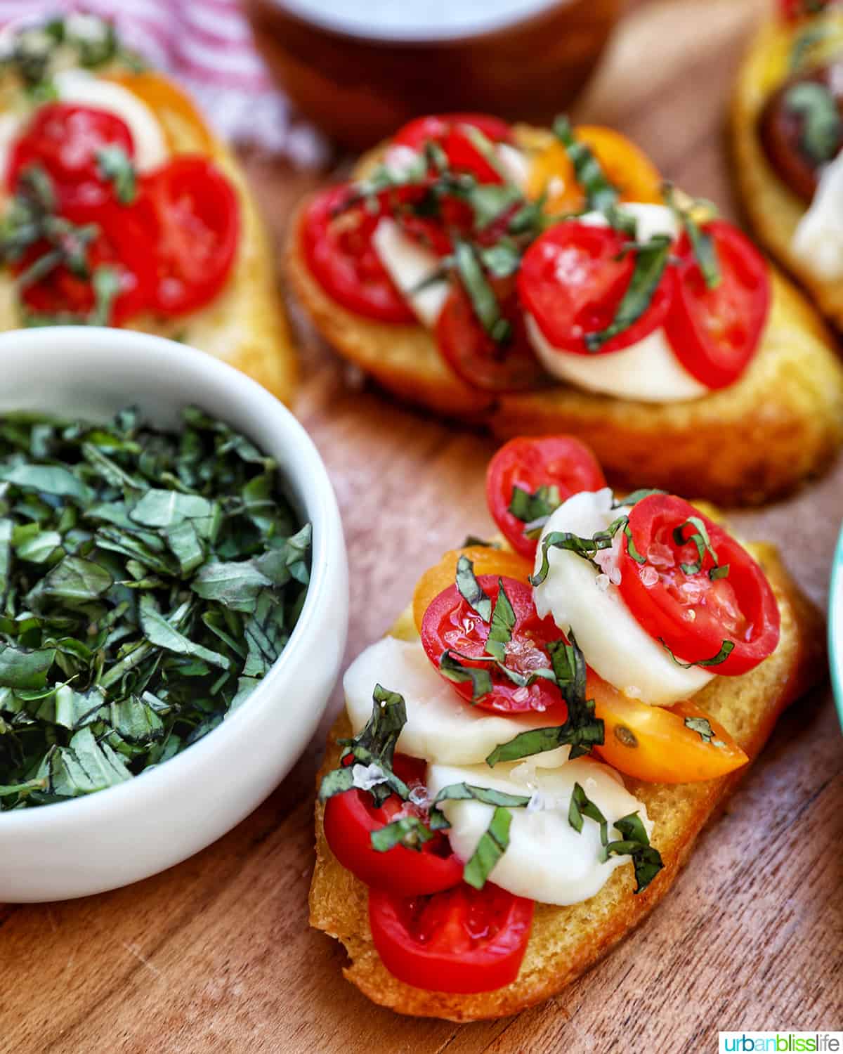 crostini with tomatoes, mozzarella, and basil on a wooden board with side of a bowl of basil.
