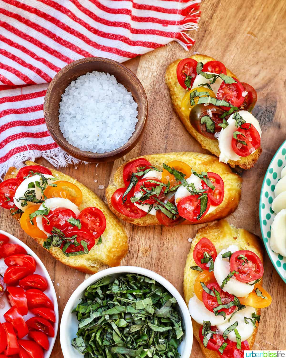 crostini with tomatoes, mozzarella, basil surrounded by plates of the same toppings and a bowl of sea salt.