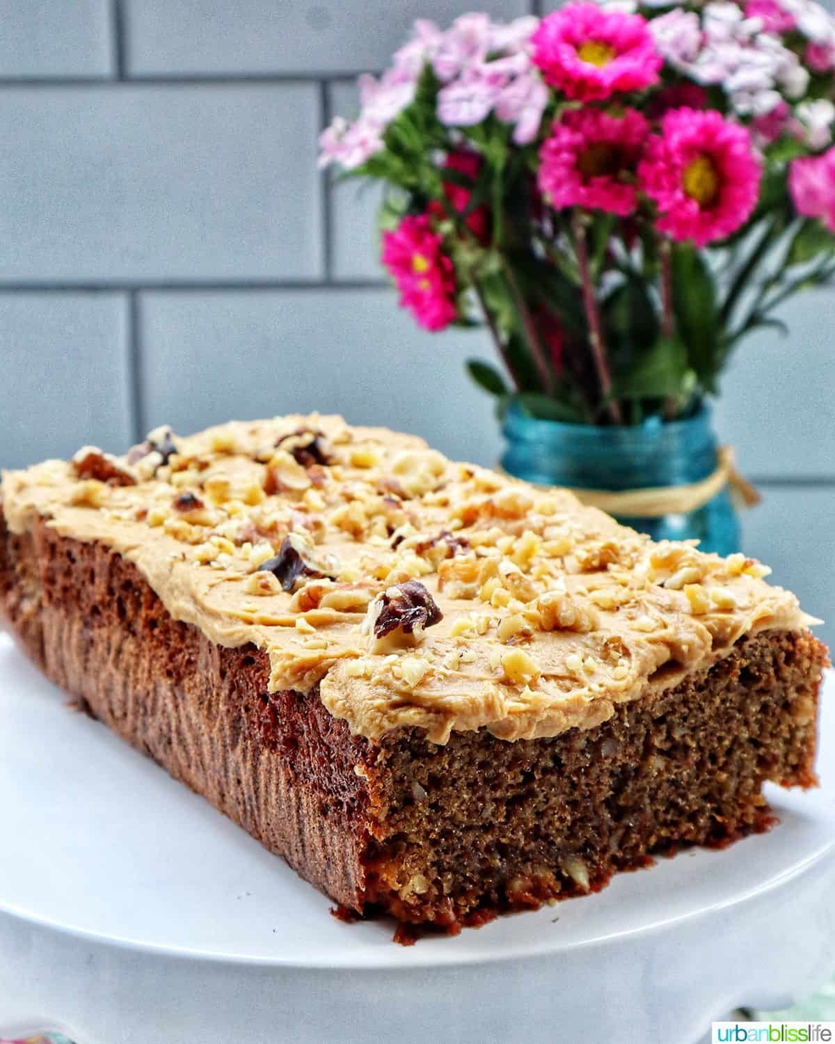 coffee walnut loaf on a white cake pedestal in front of a bouquet of spring flowers.