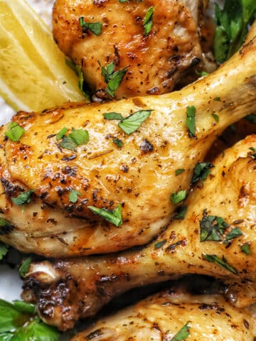 Stack of chicken drumsticks with seasoning and garnished with parsley and side of lemon wedges on bed of herbs.