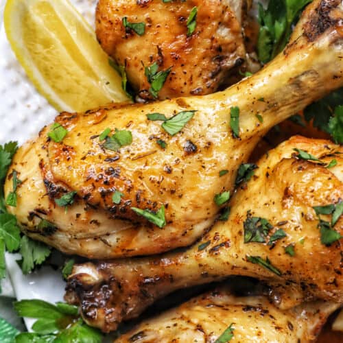 Stack of chicken drumsticks with seasoning and garnished with parsley and side of lemon wedges on bed of herbs.